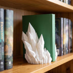Weiße Kristall-Buchstütze mit spitzen, facettierten Kristallzacken steht auf einem Holzregal vor einem aufrecht stehenden grünen Buch, links und rechts weitere Bücher mit bunten Buchrücken.