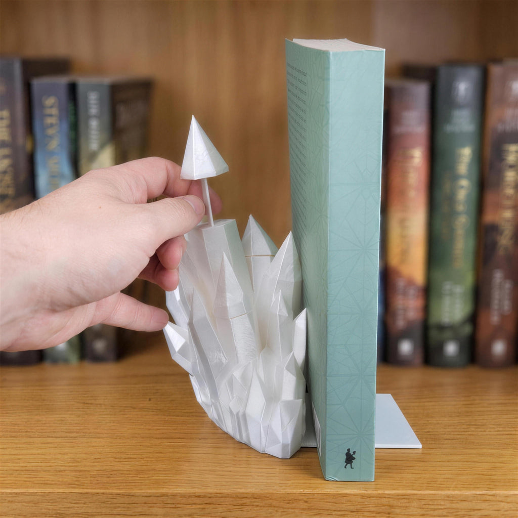Eine Hand hebt die oberste Kristallspitze mit Steckstift ab, die als separates Lesezeichen aus der weißen Kristall-Buchstütze herausgezogen wird, daneben steht ein mintgrünes Buch auf einem Holzregal.