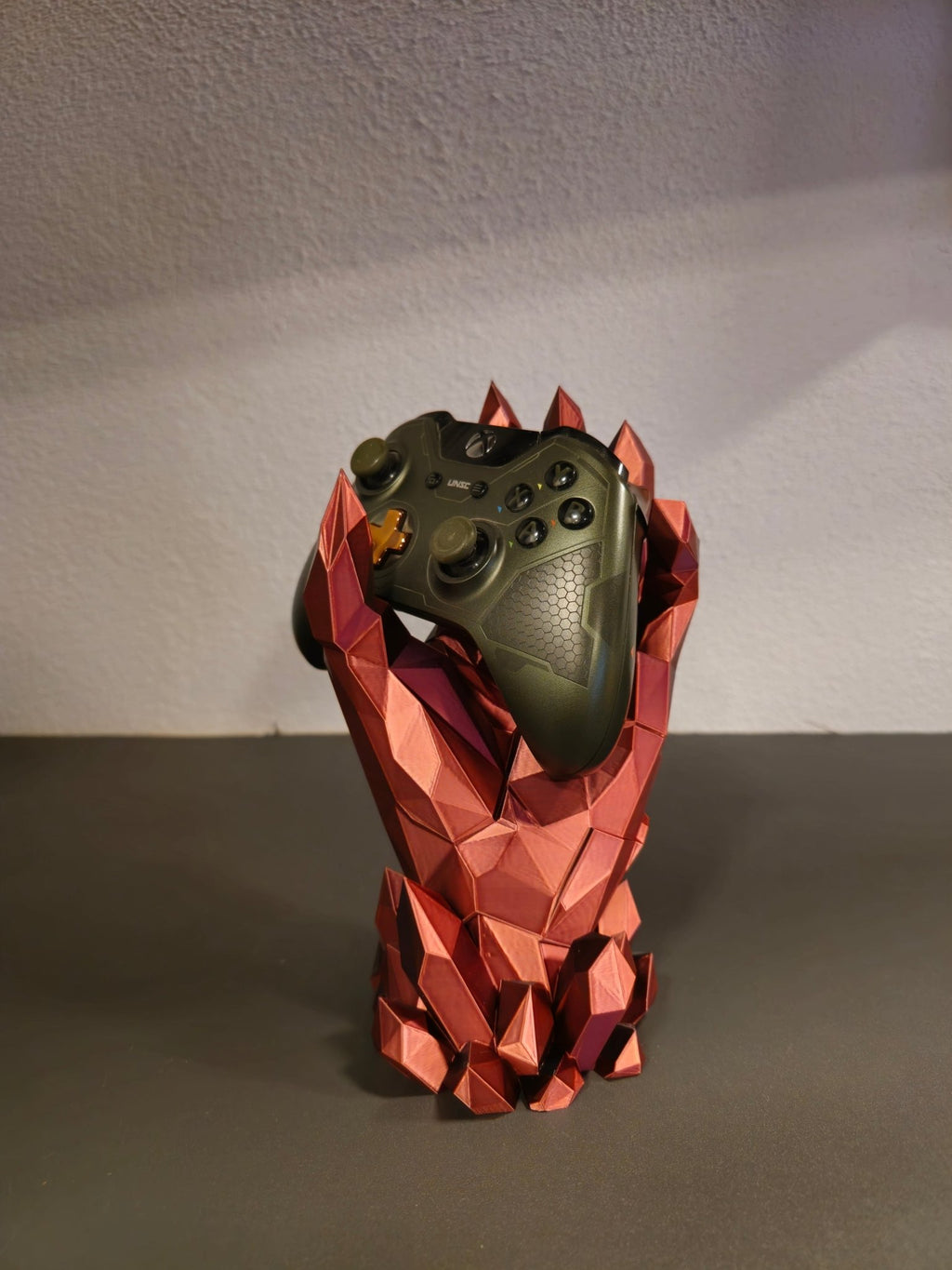 Kristallhand-Controllerhalter in Kupfer-Rot mit eingesetztem olivgrünem Gaming-Controller, Frontalansicht vor heller Wand
