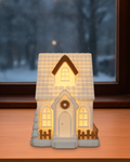 Leuchtendes Giebelhaus mit goldener Dachgaube, Kranz an der Tür und goldenem Zaun, von innen warm beleuchtet, auf einer Fensterbank vor einem Fenster mit Schneefall am Abend
