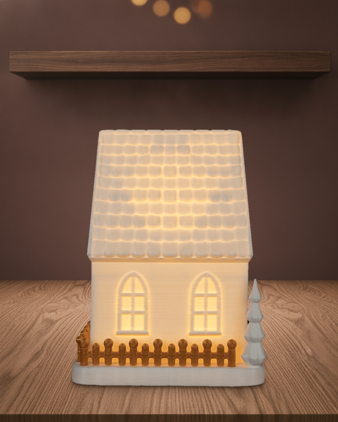 Giebelhaus von der Seite mit goldenem Zaun und warm leuchtenden Fenstern, auf einem Holztisch vor einer braunen Wand mit Regalbrett