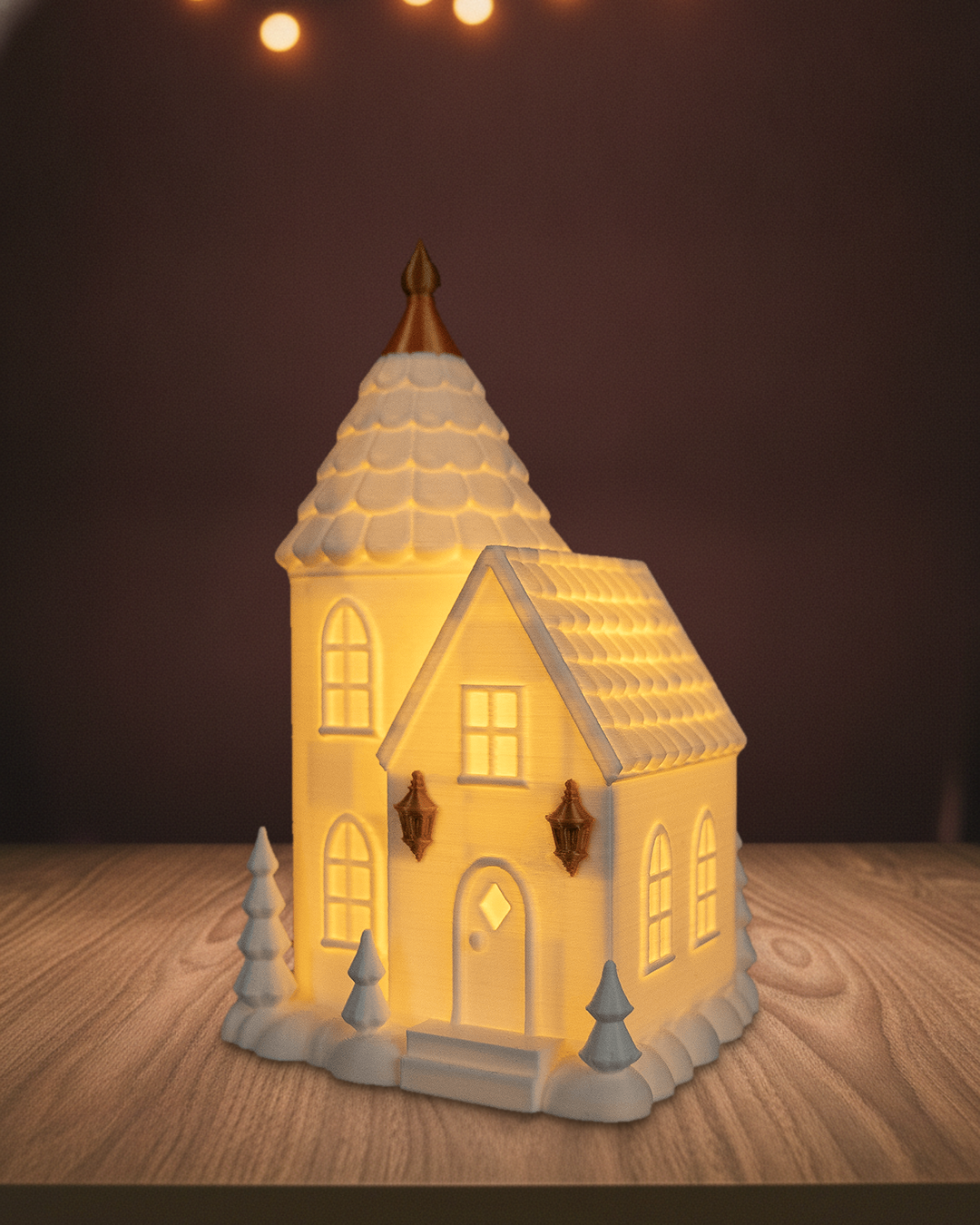 Leuchtendes Turmhaus mit Rundturm, goldener Spitze und zwei goldenen Laternen, warmes Licht in allen Fenstern, auf einem Holztisch vor einer braunen Wand mit Lichterketten
