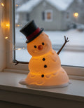 LED-Tischlampe Schneemann mit Zylinderhut und warmem Innenlicht, Frontalansicht mit sauberem Freisteller und weichem Kontaktschatten, auf weißem Fensterbrett vor winterlicher Abenddämmerung mit warmer Lichterkette
