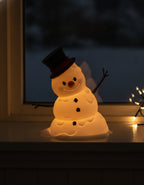 LED-Tischlampe Schneemann bei Nacht, warmes Innenlicht leuchtet durch den Körper und wirft einen sanften Lichtschein auf das Fensterbrett, dunkles verschneites Fenster im Hintergrund