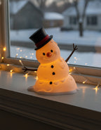 LED-Tischlampe Schneemann mit warmem Innenlicht, weiter Bildausschnitt mit mehr Fensterbrett und winterlicher Straße im Hintergrund, warme Lichterkette am Fensterrahmen