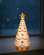 Leuchtender Weihnachtsbaum im Ornament-Design mit Spitzenmuster, goldenem Stern und goldenem Sockel, von innen warm beleuchtet, auf einer dunklen Fensterbank vor einem Fenster in der blauen Stunde