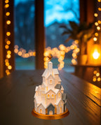 LED Winterdorf Tischlampe Turm mit beleuchteten Miniaturhäusern und goldenem Stern, Frontalansicht (weitere Aufnahme), auf dunkler Holzkonsole am Fenster in der Dämmerung, warme Lichterkette im Hintergrund