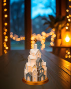 LED Winterdorf Tischlampe Turm mit beleuchteten Miniaturhäusern und goldenem Stern, Frontalansicht, auf dunkler Holzkonsole am Fenster in der Dämmerung, warme Lichterkette im Hintergrund