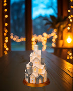 LED Winterdorf Tischlampe Turm mit beleuchteten Miniaturhäusern und goldenem Stern, leicht gedrehte Ansicht, auf dunkler Holzkonsole am Fenster in der Dämmerung, warme Lichterkette im Hintergrund
