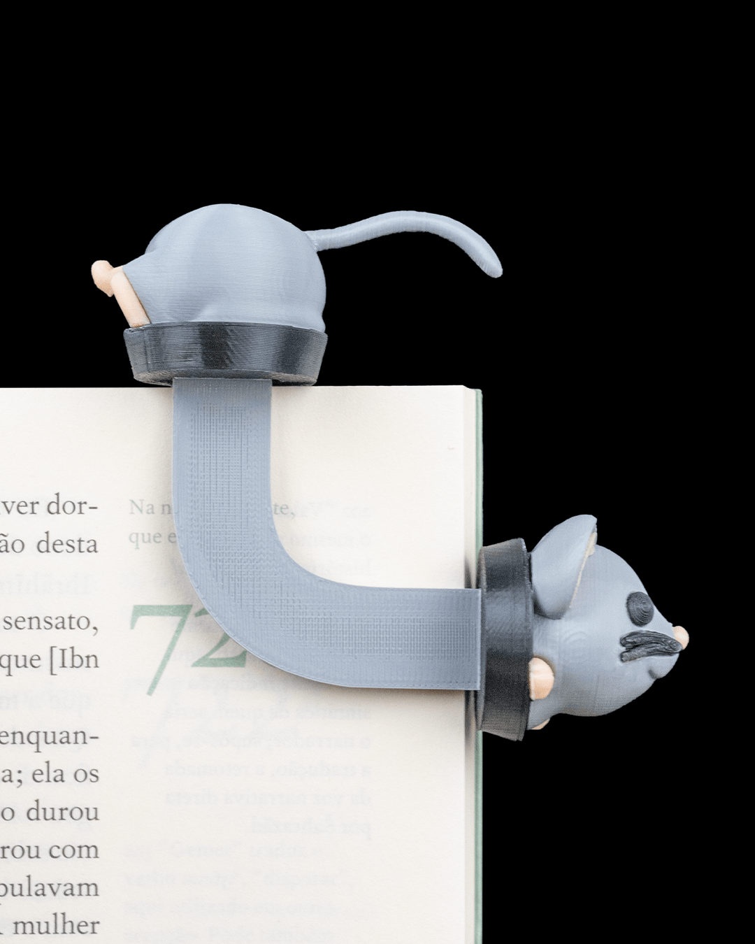 Graues Maus-Lesezeichen auf aufgeschlagenem Buch mit lesbarem Text, Rute oben auf der Buchkante, Kopf mit Schnurrhaaren seitlich unter der Seite hervorschauend
