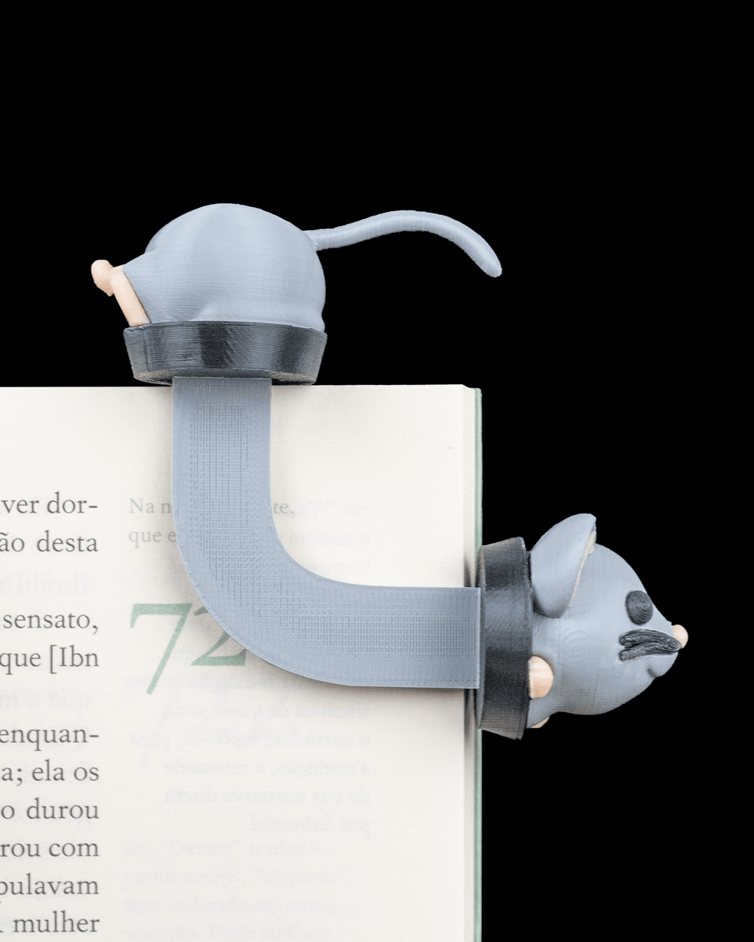 Graues Maus-Lesezeichen aus dem 3D-Drucker auf aufgeschlagenem Buch mit lesbarem Text – geringelter Schwanz ragt oben aus den Seiten, Kopf mit rosa Ohren und Schnurrhaaren schaut seitlich über die Buchkante