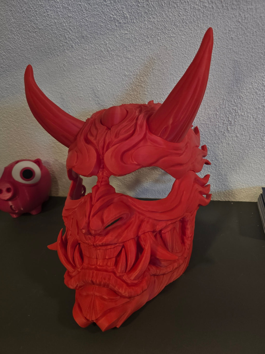 Oni-Maske Der Dämon in Rot, Frontalansicht schräg von links auf dunklem Regal, Deko-Figuren im Hintergrund – Münch3D