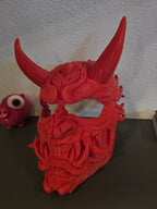 Oni-Maske Der Dämon in Rot, Frontalansicht schräg von links auf dunklem Regal, Deko-Figuren im Hintergrund – Münch3D