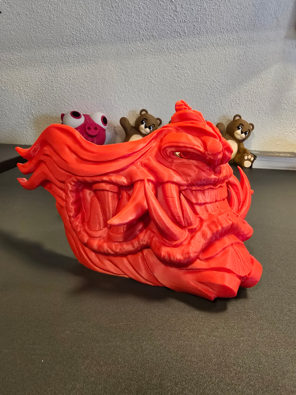 Oni-Maske Der Dämon in Rot, seitliche Nahaufnahme des geöffneten Mauls, Deko-Figuren im Hintergrund – Münch3D