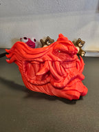 Oni-Maske Der Dämon in Rot, seitliche Nahaufnahme des geöffneten Mauls, Deko-Figuren im Hintergrund – Münch3D