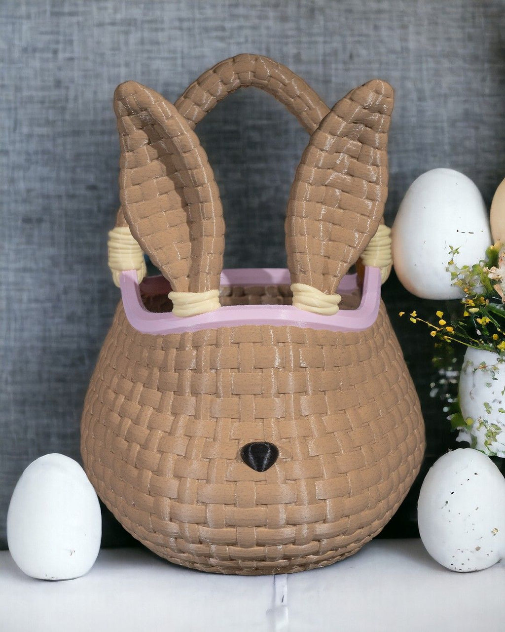 Geflochtenes XL Osterkörbchen in Hasenform mit spitzen Ohren, geflochtenem Henkel und rosa Randfarbe, vor grauem Stoffhintergrund mit weißen Deko-Eiern und gelben Blüten.