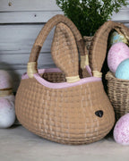 Geflochtenes XL Osterkörbchen in Hasenform mit spitzen Ohren, geflochtenem Henkel und rosa Randfarbe, vor weißer Holzverschalung mit grüner Pflanze, Weidenkorb mit pastellfarbenen Eiern und rosa-blau gesprenkelten Ostereiern.