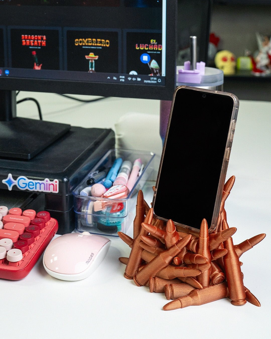 Kupferfarbener Patronen-Handyhalter mit eingestecktem Smartphone in Hülle auf Gaming-Schreibtisch, Monitor und Deko im Hintergrund