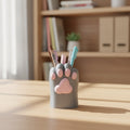 Stiftehalter in Katzenpfotenform in Grau mit rosa Ballen, mit bunten Stiften befüllt, auf hellem Holzschreibtisch mit Bücherregal im Hintergrund