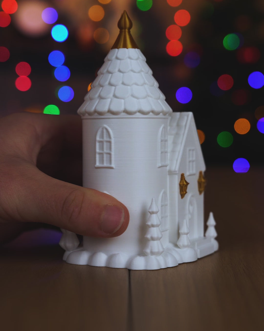 Kurzvideo: Giebelhaus und Turmhaus in Weiß und Gold werden in der Hand gedreht und gezeigt, im Hintergrund Weihnachtsbaum-Lichter, im Dunkeln scheint warmes Licht durch die Fenster
