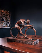 Handbemalte Radfahrer-Statue in Kupfer-Bronze-Optik – Lifestyle-Aufnahme auf Holztisch vor dunkler Wand mit Pinnwand