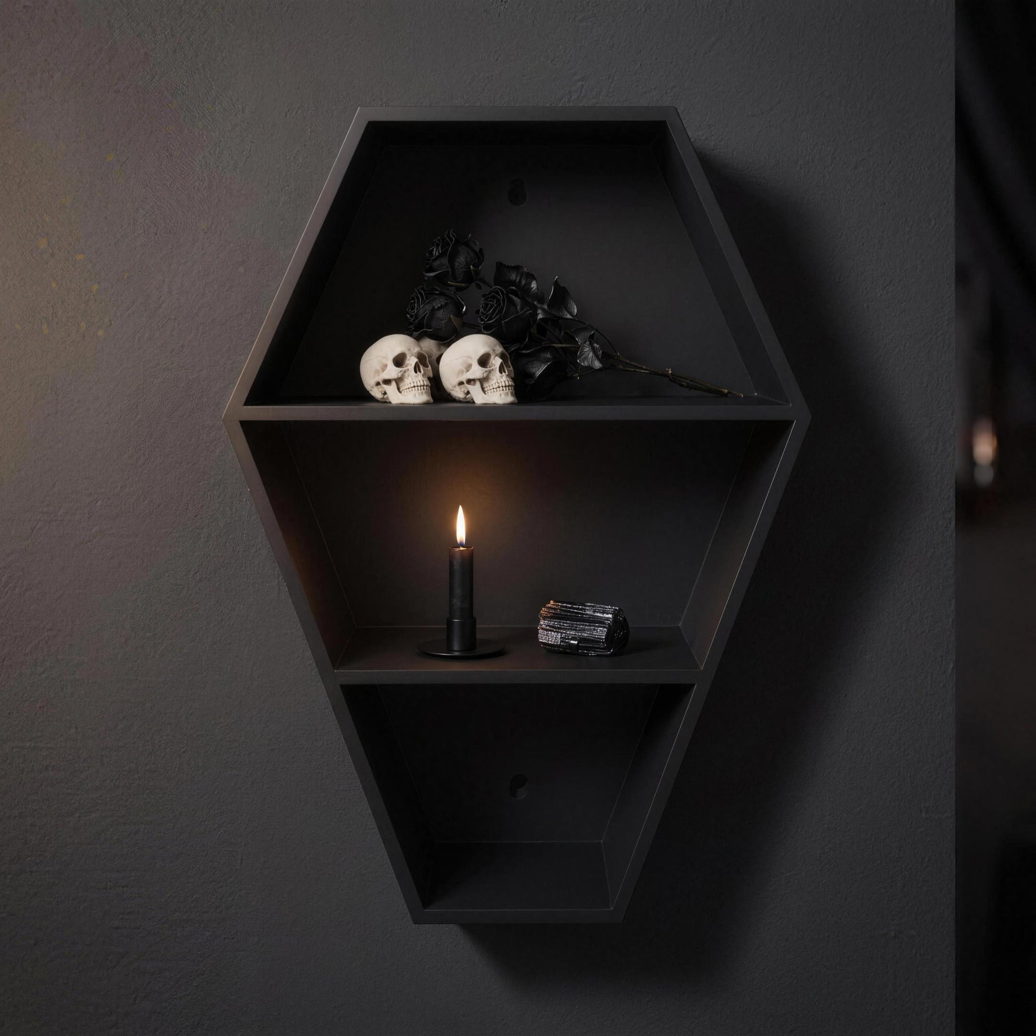 Matte black coffin shelf with two skulls, black roses, a lit candle and a black crystal on a dark wall