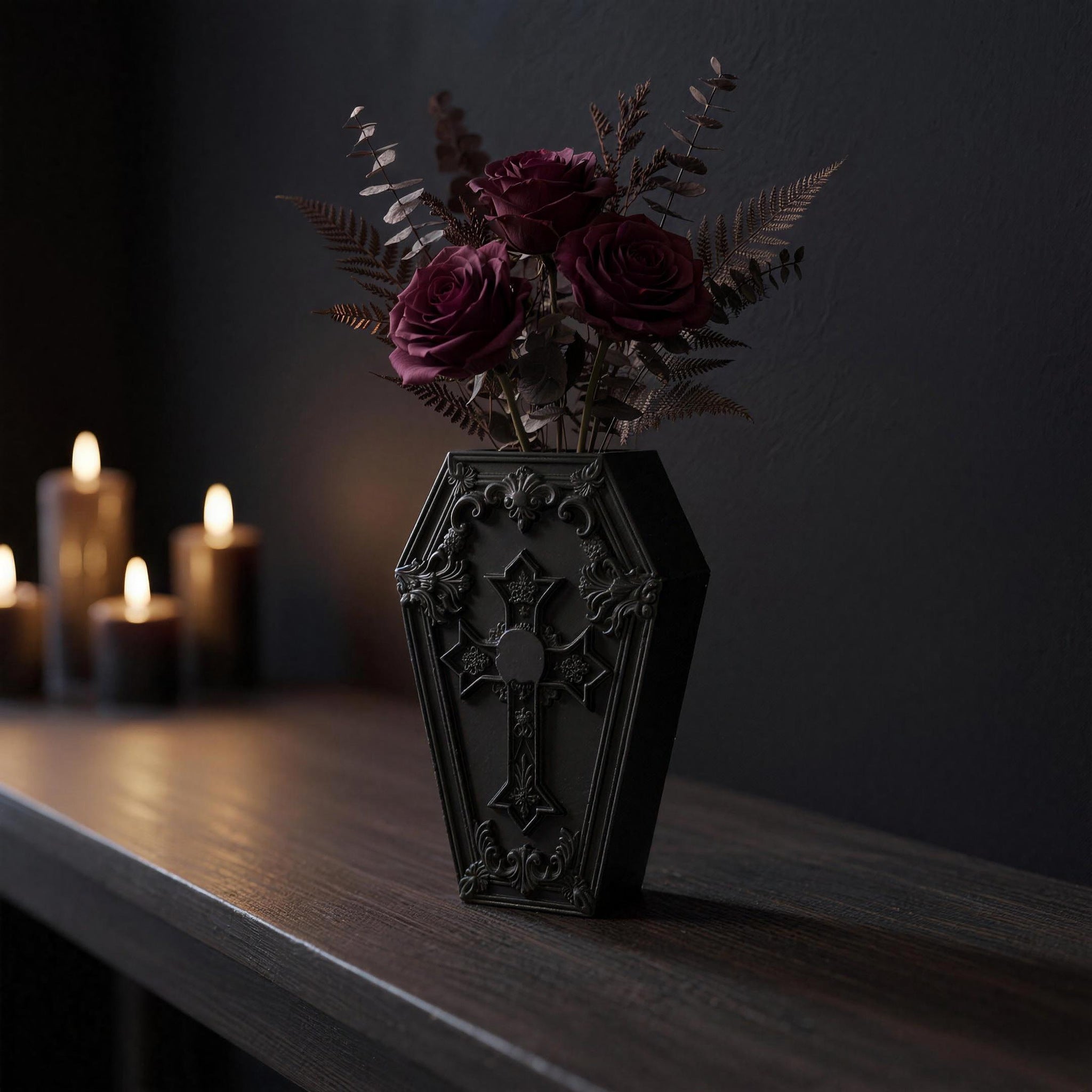 Black gothic coffin vase with cross-and-skull relief and dark red roses on a dark sideboard, front view with candlelight in the background