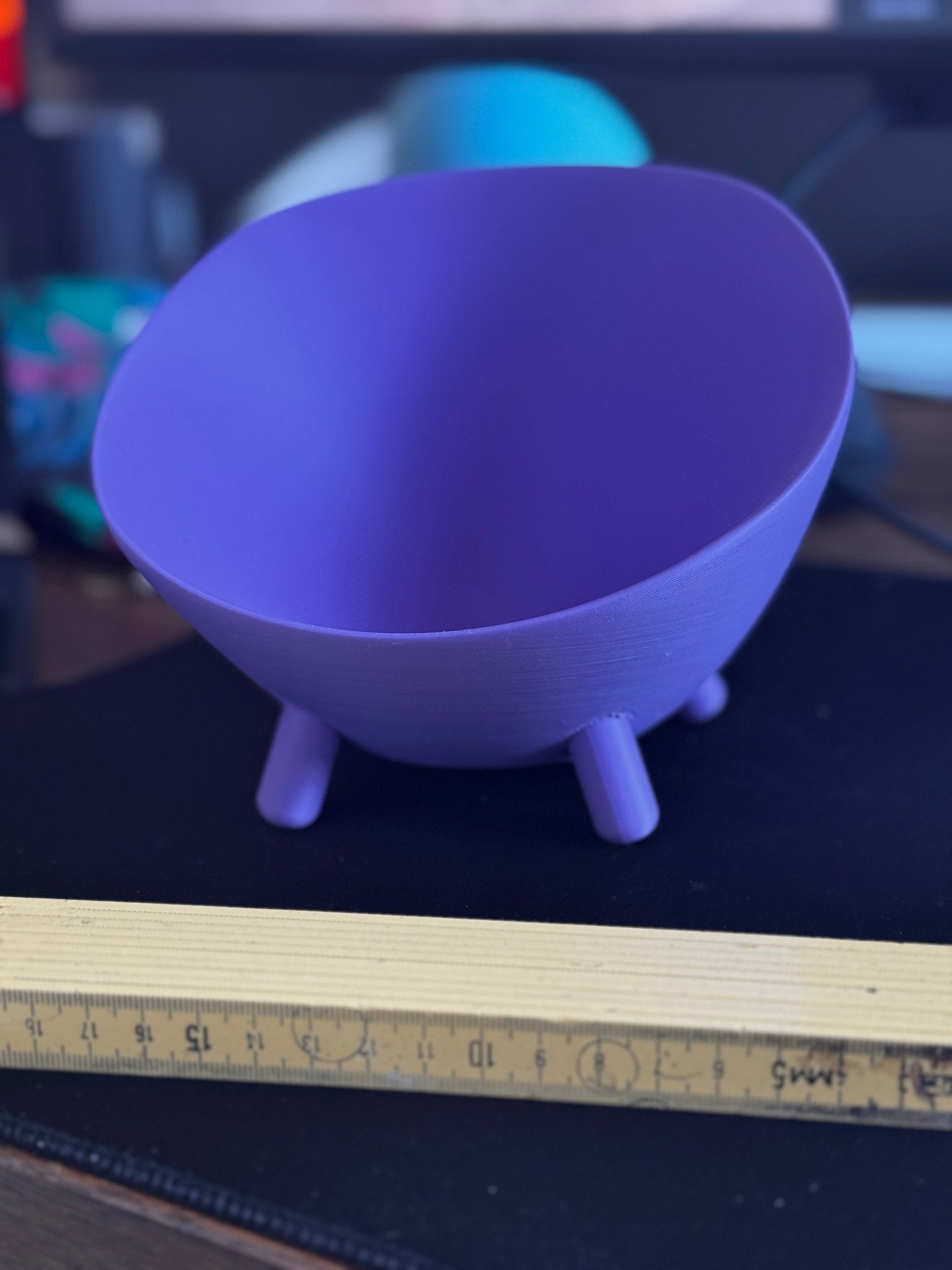 Lila Schale mit Füßen neben ausgeklapptem Zollstock zum Größenvergleich, auf dunklem Schreibtisch, im Hintergrund unscharfes blaues Licht und Tastatur.
