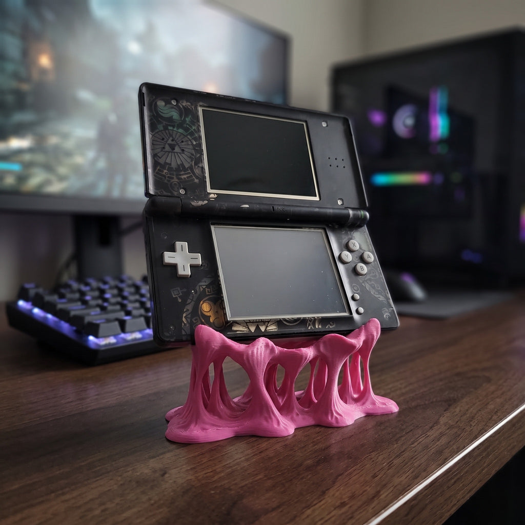 Pinker Tropfen-Konsolenhalter aus 3D-Druck mit organisch geformten, durchbrochenen Beinen, darauf eine aufgeklappte Handheld-Spielkonsole mit Doppelbildschirm, im Hintergrund ein Gaming-Setup mit beleuchteter Tastatur und Monitoren
