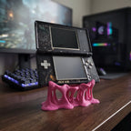 Pinker Tropfen-Konsolenhalter aus 3D-Druck mit organisch geformten, durchbrochenen Beinen, darauf eine aufgeklappte Handheld-Spielkonsole mit Doppelbildschirm, im Hintergrund ein Gaming-Setup mit beleuchteter Tastatur und Monitoren