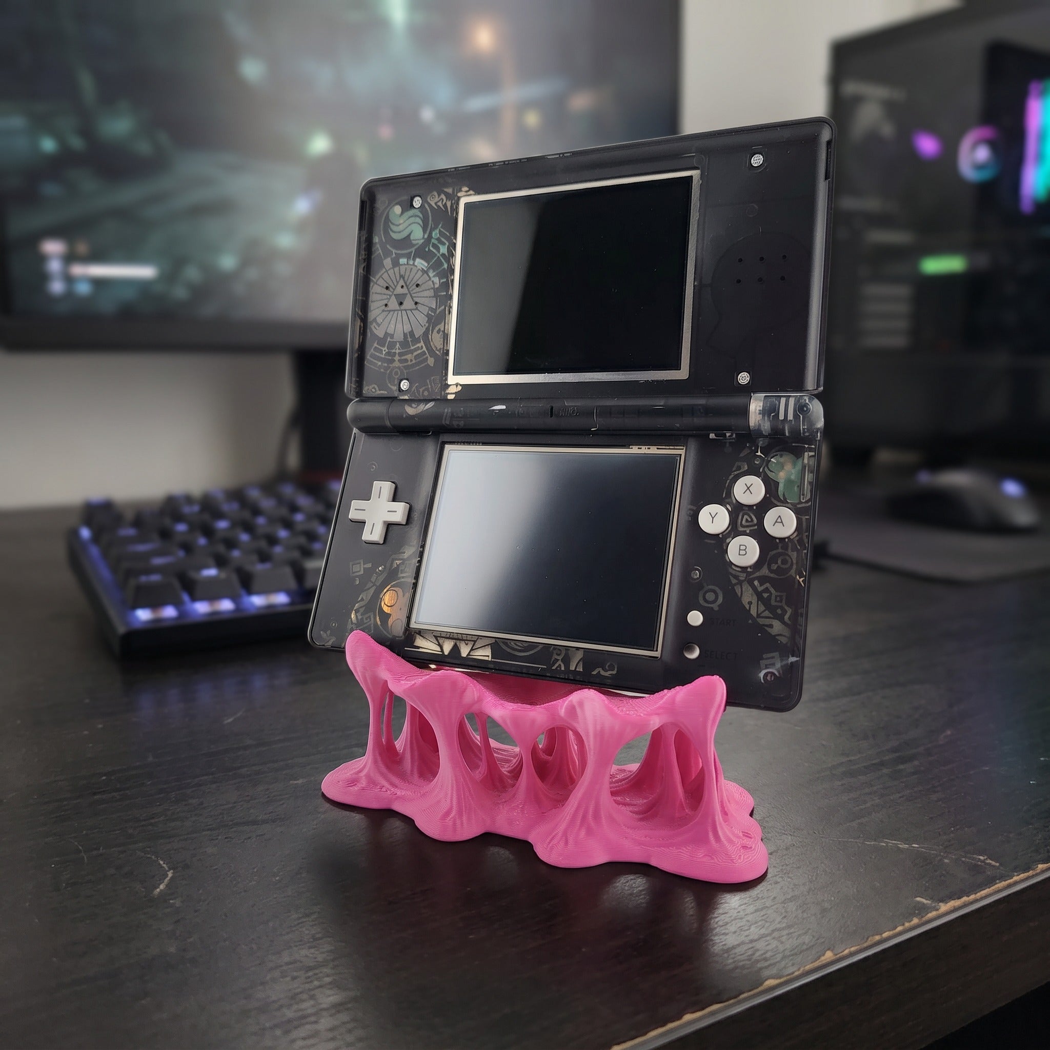 Zentrale Frontansicht des pinken Tropfen-Konsolenhalters aus 3D-Druck mit geschmolzen wirkenden, tropfenförmigen Beinen, darauf eine aufklappbare Handheld-Spielkonsole mit zwei Bildschirmen und Steuerkreuz auf einem dunklen Schreibtisch