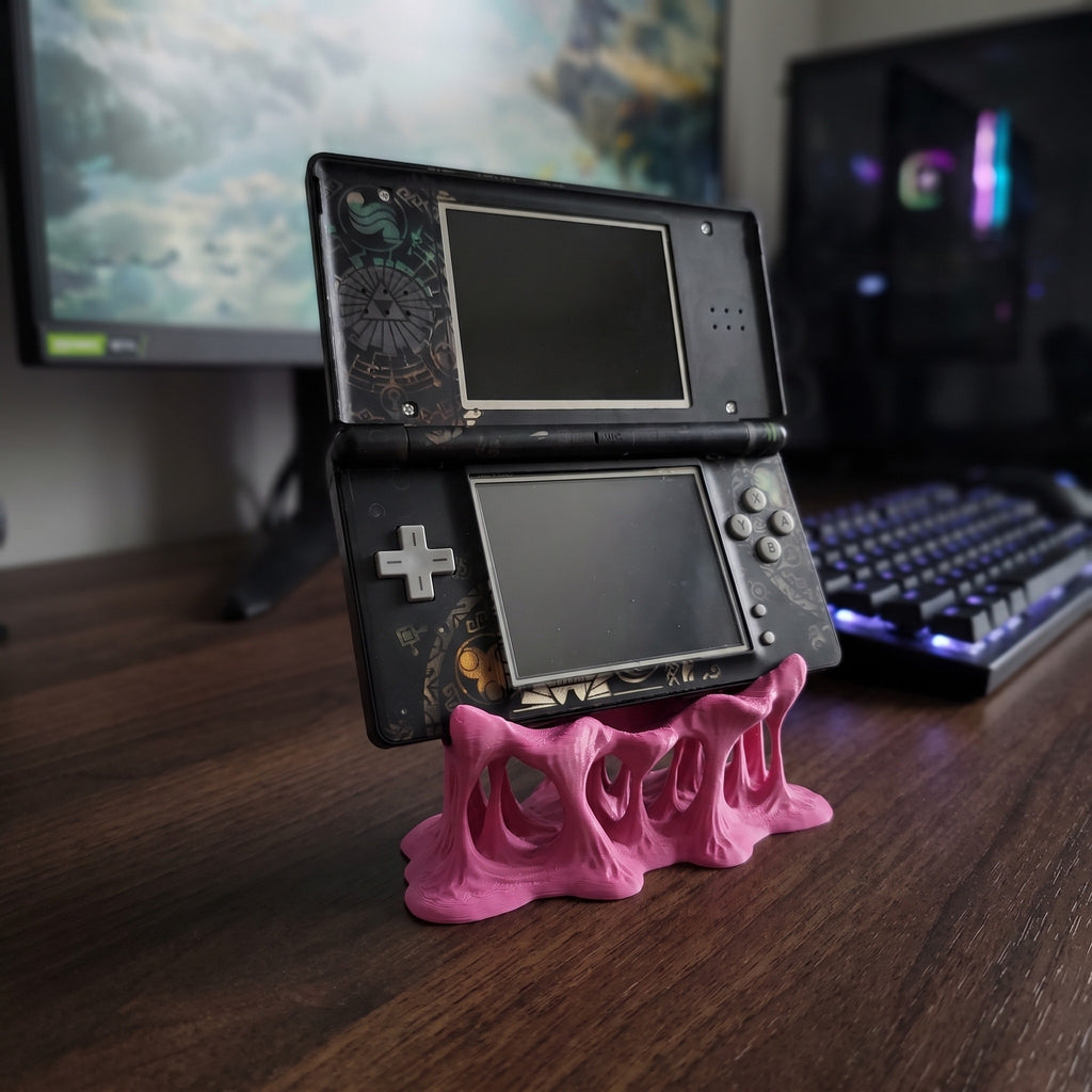 Leicht seitliche Ansicht des pinken Tropfen-Konsolenhalters mit tropfenförmigen, ausgehöhlten Beinen im Schmelz-Look, darauf eine aufklappbare Handheld-Spielkonsole mit Doppelbildschirm auf einem Holzschreibtisch