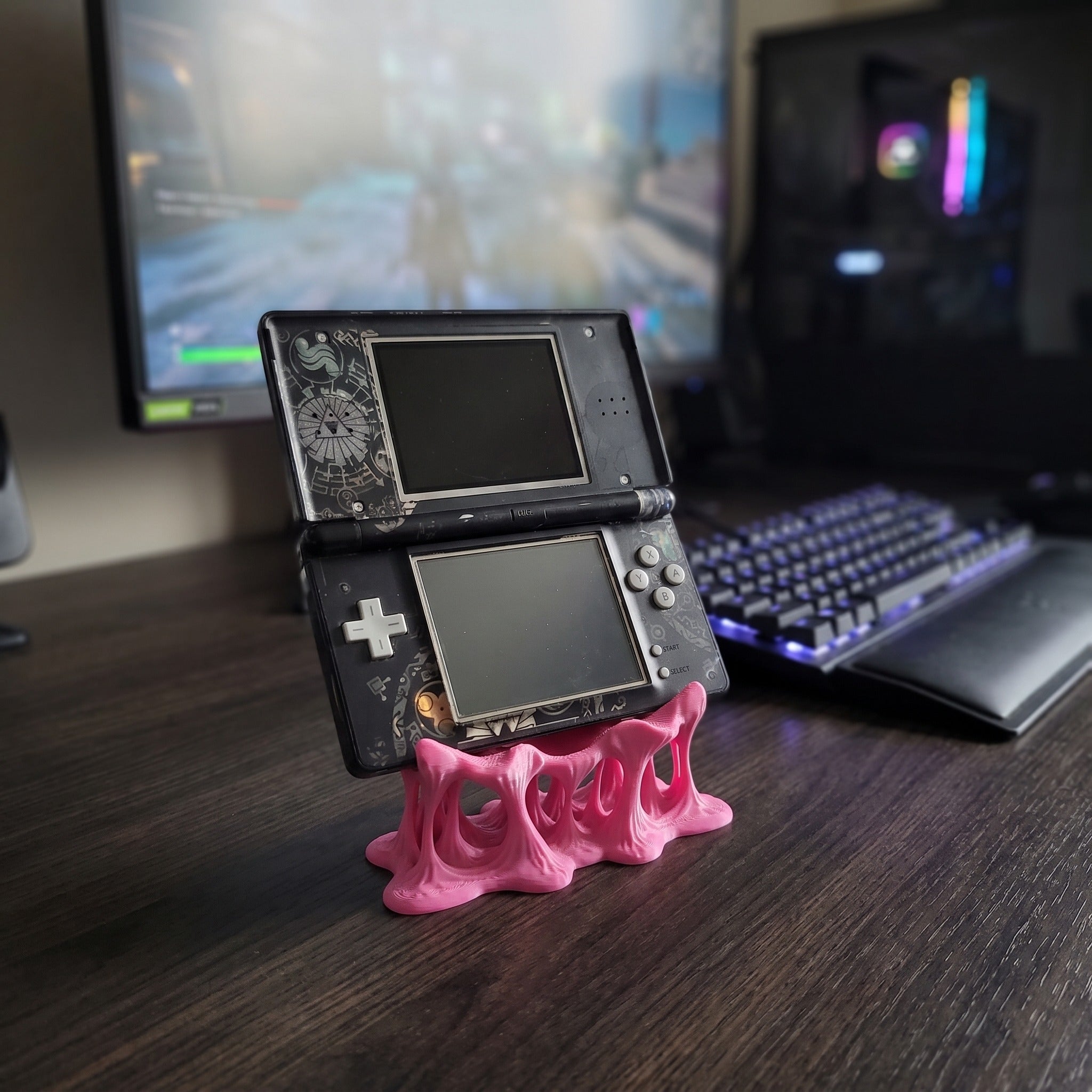 Nahaufnahme des pinken Tropfen-Konsolenhalters aus 3D-Druck mit Steuerkreuz und Actionknöpfen der aufgestellten Handheld-Spielkonsole im Vordergrund, dahinter eine beleuchtete Gaming-Tastatur