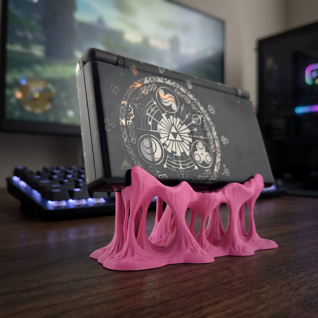 Seitliche Rückansicht des pinken Tropfen-Konsolenhalters mit mehreren geschmolzen wirkenden Beinen, darauf eine aufklappbare Handheld-Spielkonsole mit rundem Grafik-Aufkleber auf der Rückseite