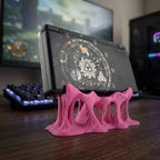 Seitliche Rückansicht des pinken Tropfen-Konsolenhalters mit mehreren geschmolzen wirkenden Beinen, darauf eine aufklappbare Handheld-Spielkonsole mit rundem Grafik-Aufkleber auf der Rückseite