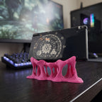 Rückansicht des pinken Tropfen-Konsolenhalters mit mehreren durchbrochenen, tropfenartigen Beinen im Schmelz-Look, darauf eine aufklappbare Handheld-Spielkonsole mit rundem Grafik-Aufkleber, im Hintergrund Gaming-Monitore