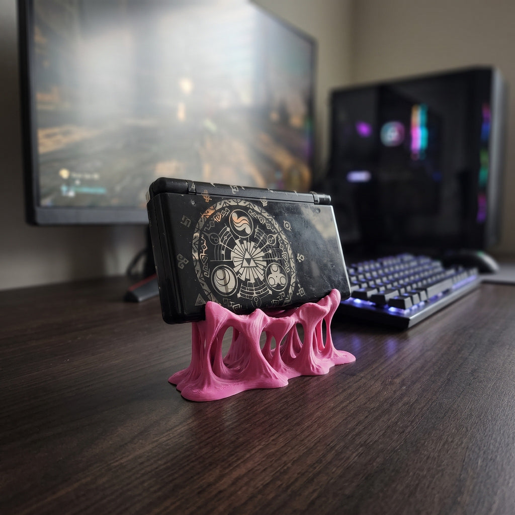 Weitere Rückansicht des pinken Tropfen-Konsolenhalters aus 3D-Druck auf einem dunklen Schreibtisch, darauf eine aufklappbare Handheld-Spielkonsole mit rundem Grafik-Aufkleber, im Hintergrund eine beleuchtete Tastatur und Monitore
