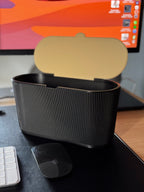 Derselbe schwarze geriffelte Mülleimer mit geöffnetem Schwingdeckel, Innenseite des Deckels in Gold/Beige sichtbar, leer, auf demselben Schreibtisch mit iMac im Hintergrund.