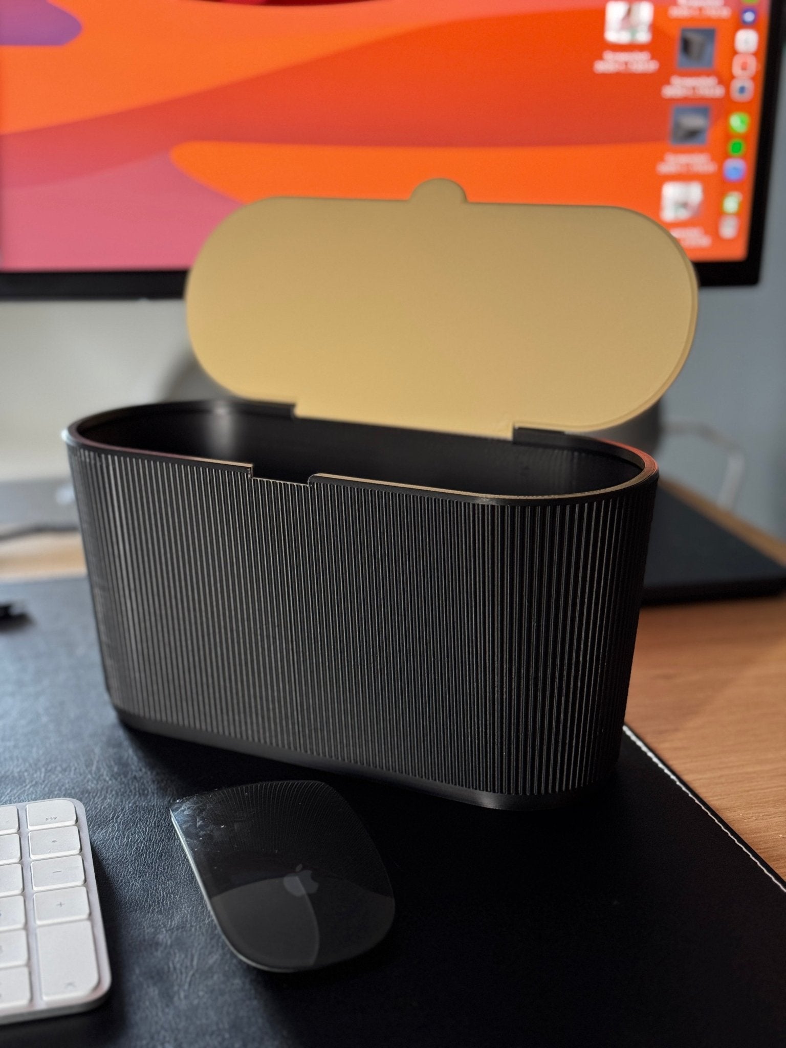 Derselbe schwarze geriffelte Mülleimer mit geöffnetem Schwingdeckel, Innenseite des Deckels in Gold/Beige sichtbar, leer, auf demselben Schreibtisch mit iMac im Hintergrund.