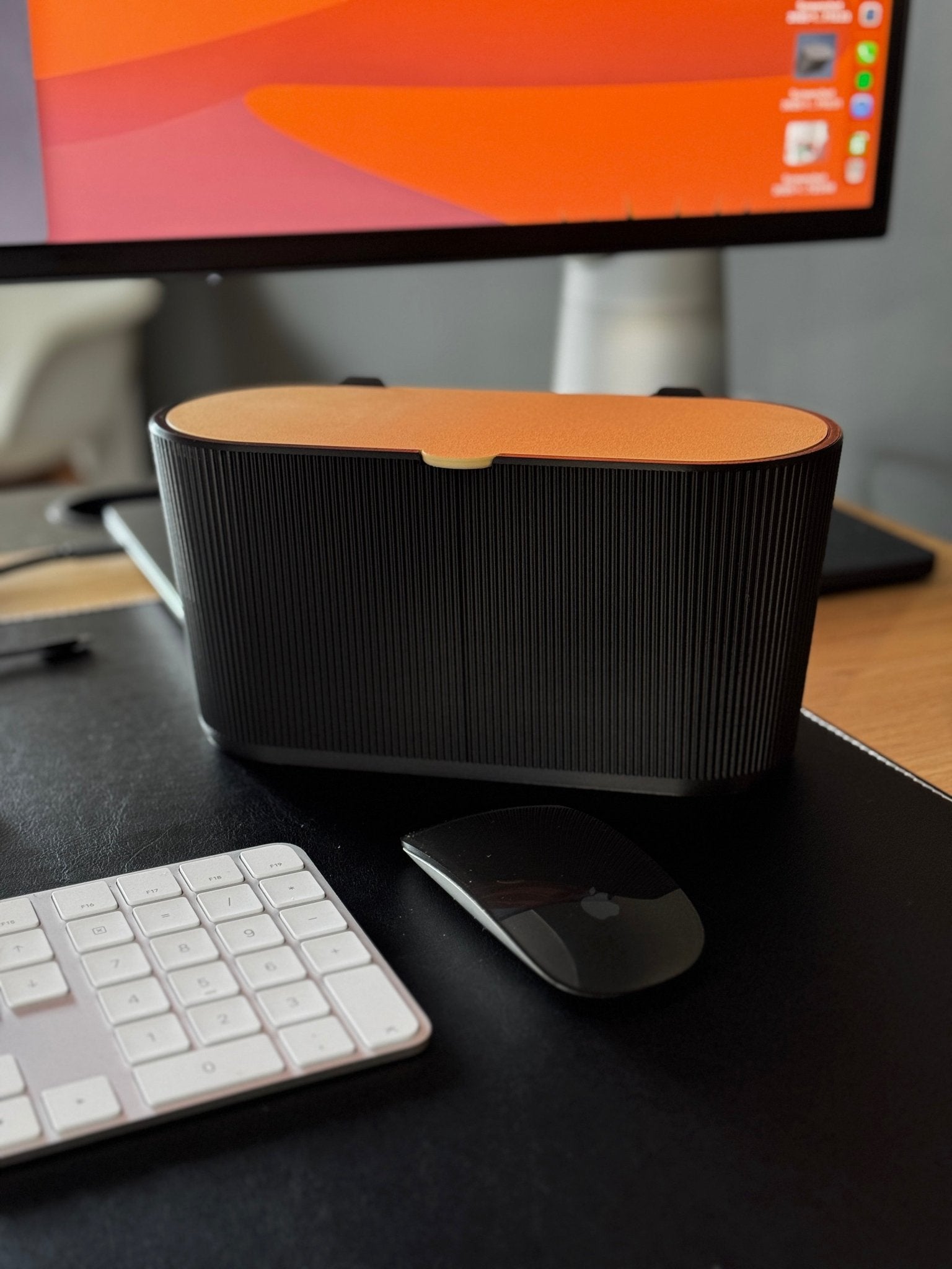 Geriffelter ovaler Schreibtisch-Mülleimer in Schwarz mit orangefarbenem Schwingdeckel, geschlossen auf einem echten Schreibtisch mit iMac, Apple-Tastatur und -Maus im Hintergrund.