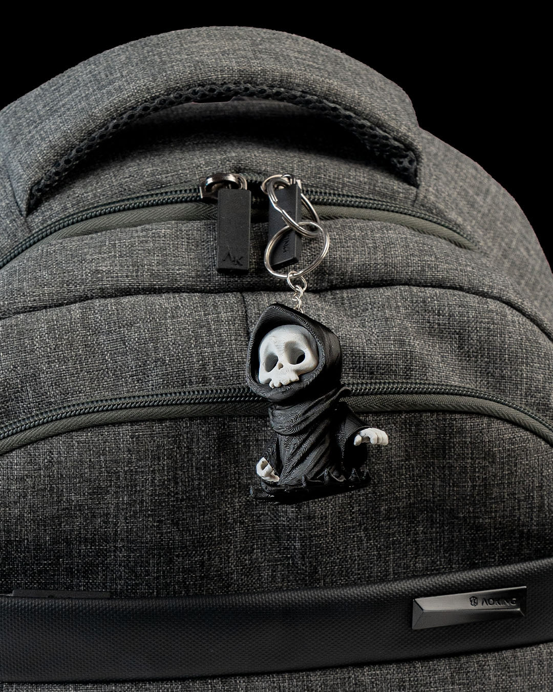 Sensenmann-Schlüsselanhänger hängt am Reißverschluss eines grauen Rucksacks, befestigt mit Metallring und Kette – zeigt die Nutzung als Taschenanhänger.