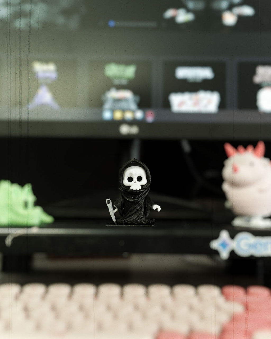 Kleine Sensenmann-Schlüsselanhänger-Figur steht vor einem Monitor auf einem Schreibtisch, umgeben von weiteren Mini-Sammelfiguren und einer Tastatur.