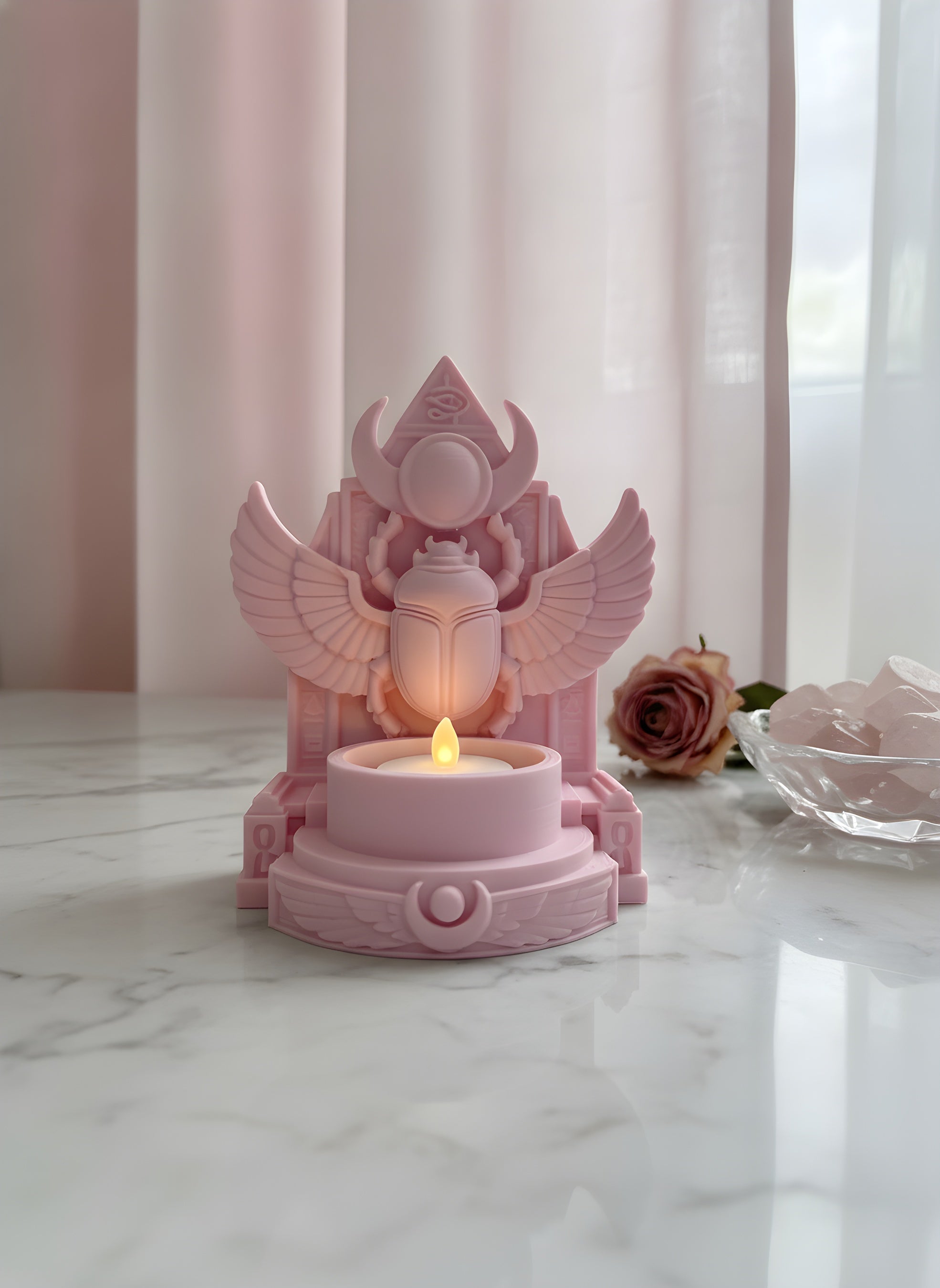 Rosa geflügelter Skarabäus-Teelichthalter vor pastellrosa Vorhang mit Rosenquarz-Schale und getrockneter Rose, LED-Teelicht-Schein