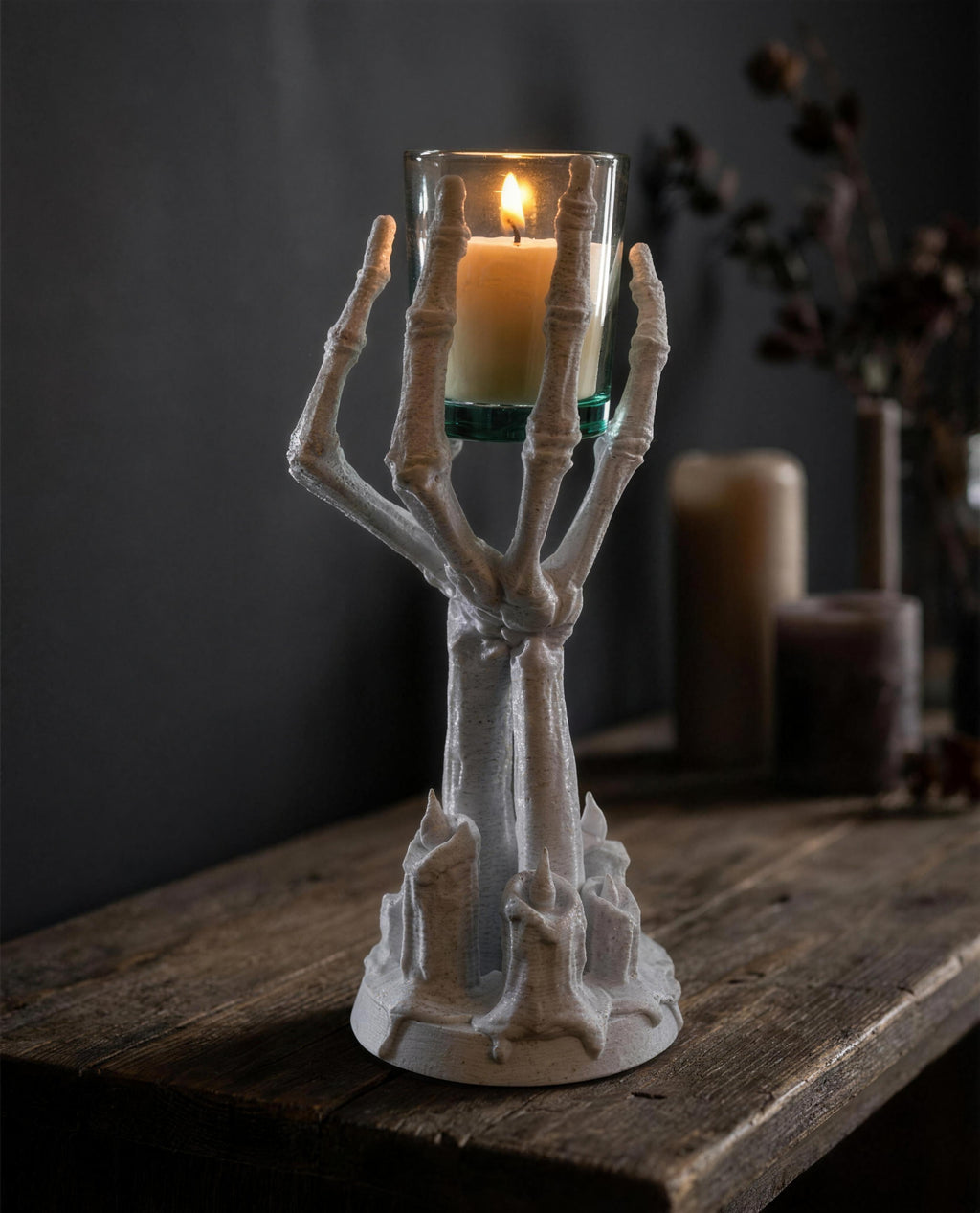3D-gedruckter Skeletthand-Kerzenhalter in Marmor-Knochen-Optik hält ein Teelichtglas mit brennender Kerze, dunkles Gothic-Sideboard im Hintergrund