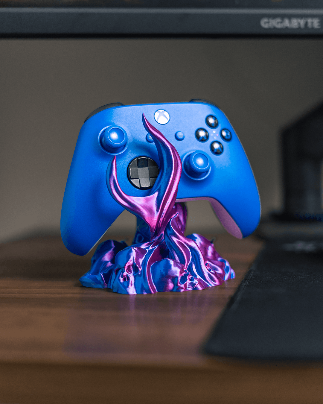 Skullflame Controller-Halter mit blauem Gaming-Controller, auf Holz-Schreibtisch mit Mauspad, im Hintergrund ein Gaming-Monitor mit Spielszene.