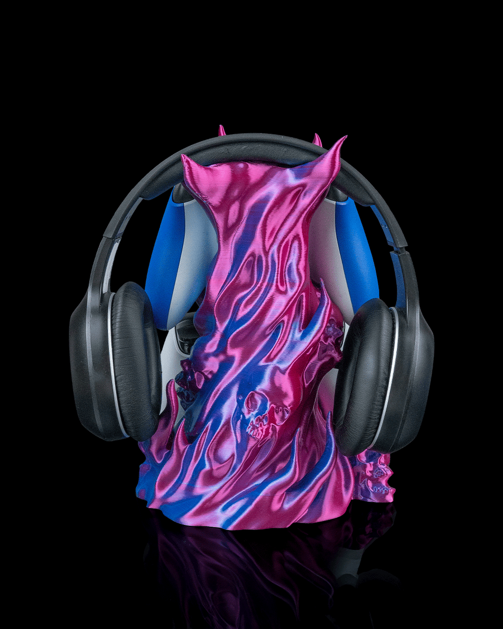 Skullflame Headset & Controller Ständer in Pink-Blau, Frontalansicht freigestellt vor schwarzem Hintergrund, mit hängendem Over-Ear-Kopfhörer, blauer und weißer Gaming-Controller teilweise hinter den Flammen sichtbar, Totenkopf-Relief mittig.