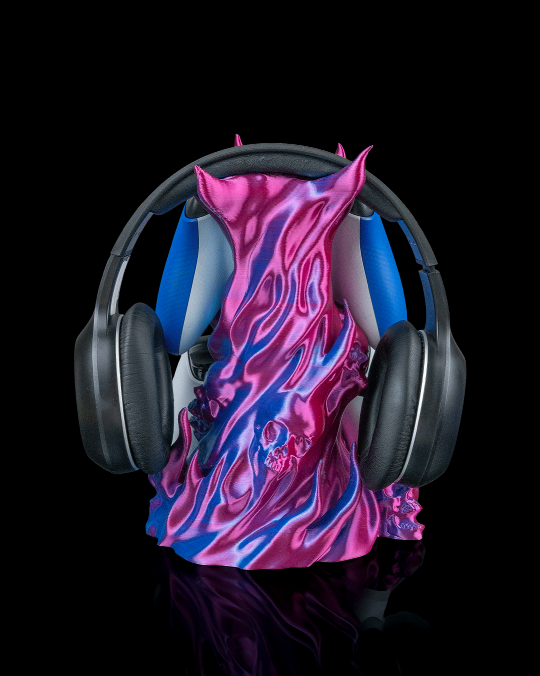 Skullflame Headset & Controller Ständer in Pink-Blau, Frontalansicht freigestellt vor schwarzem Hintergrund, mit hängendem Over-Ear-Kopfhörer, blauer und weißer Gaming-Controller teilweise hinter den Flammen sichtbar, Totenkopf-Relief mittig.
