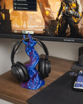 Skullflame Headset-Halter in Blau-Lila mit aufgehängtem Over-Ear-Kopfhörer, auf einem Gaming-Schreibtisch mit Mauspad und Tastatur, im Hintergrund ein Monitor mit einem Action-Spiel.