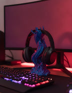 Skullflame Headset-Halter in Blau-Lila mit aufgehängtem Over-Ear-Kopfhörer, Nahaufnahme auf Gaming-Schreibtisch mit RGB-Tastatur im Vordergrund und unscharfem Monitor im Hintergrund.