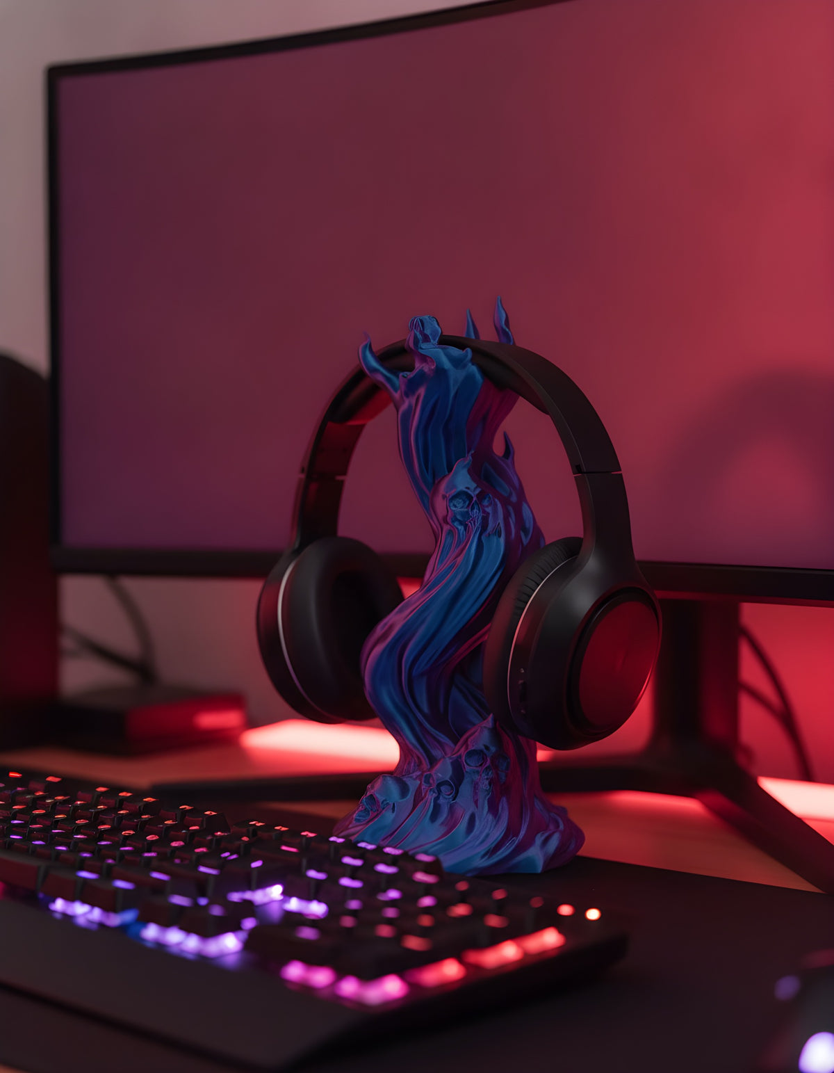 Skullflame Headset-Halter in Blau-Lila mit aufgehängtem Over-Ear-Kopfhörer, Nahaufnahme auf Gaming-Schreibtisch mit RGB-Tastatur im Vordergrund und unscharfem Monitor im Hintergrund.